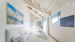 A hotel room with a large bed, a painting of a sunset, a TV, and a bathroom with a mirror and sink. - Oasi Blu Sardinia (Pula)