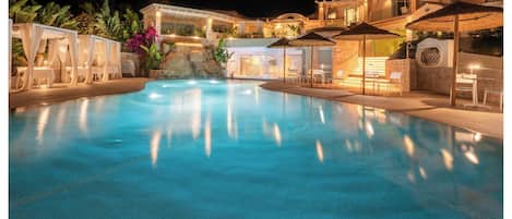 A swimming pool with a stone wall and a waterfall feature, surrounded by outdoor seating and umbrellas.