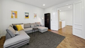 A modern living room with a grey sectional sofa, a small round coffee table, and a dark bookshelf. There are three abstract paintings on the wall and a white door leading to another room. - Big spacious central apartment in Vienna (Vienna)