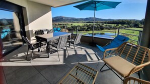 Ein Balkon mit Tisch und Stühlen, einem blauen Sonnenschirm und Blick auf eine grüne Landschaft mit Hügeln.