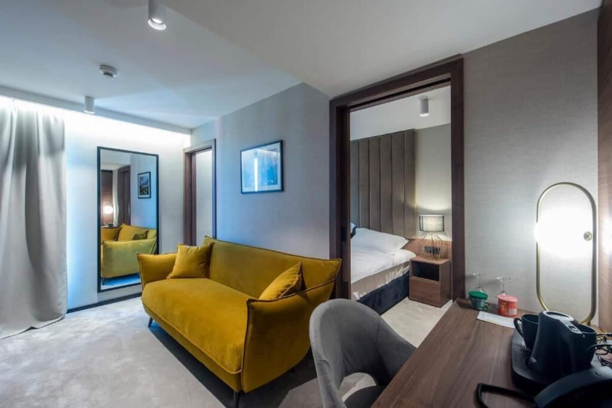 A hotel room with a yellow sofa, a bed, a desk with a lamp, and a mirror.