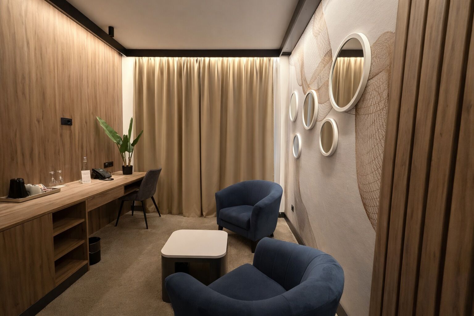 Una habitación de hotel moderna con un escritorio de madera, dos sillones azules, una mesita y una pared con espejos circulares.