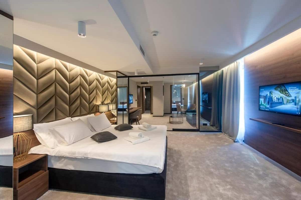 A modern hotel room with a large bed, a TV mounted on the wall, and a view of the city through a glass partition.