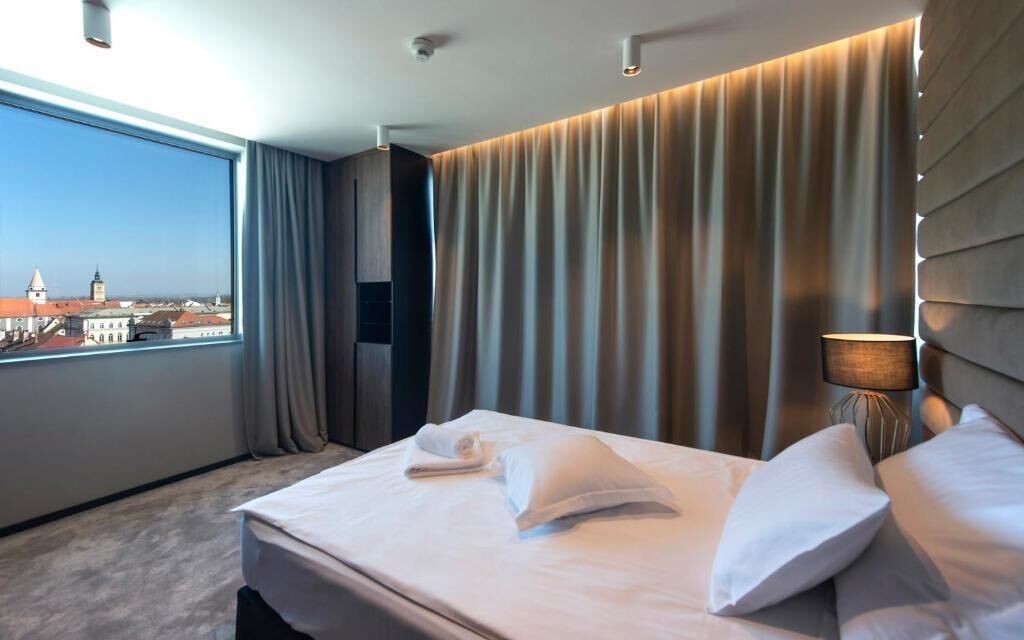 A hotel room with a large window, a bed with white linens, a city view, and a modern lamp.