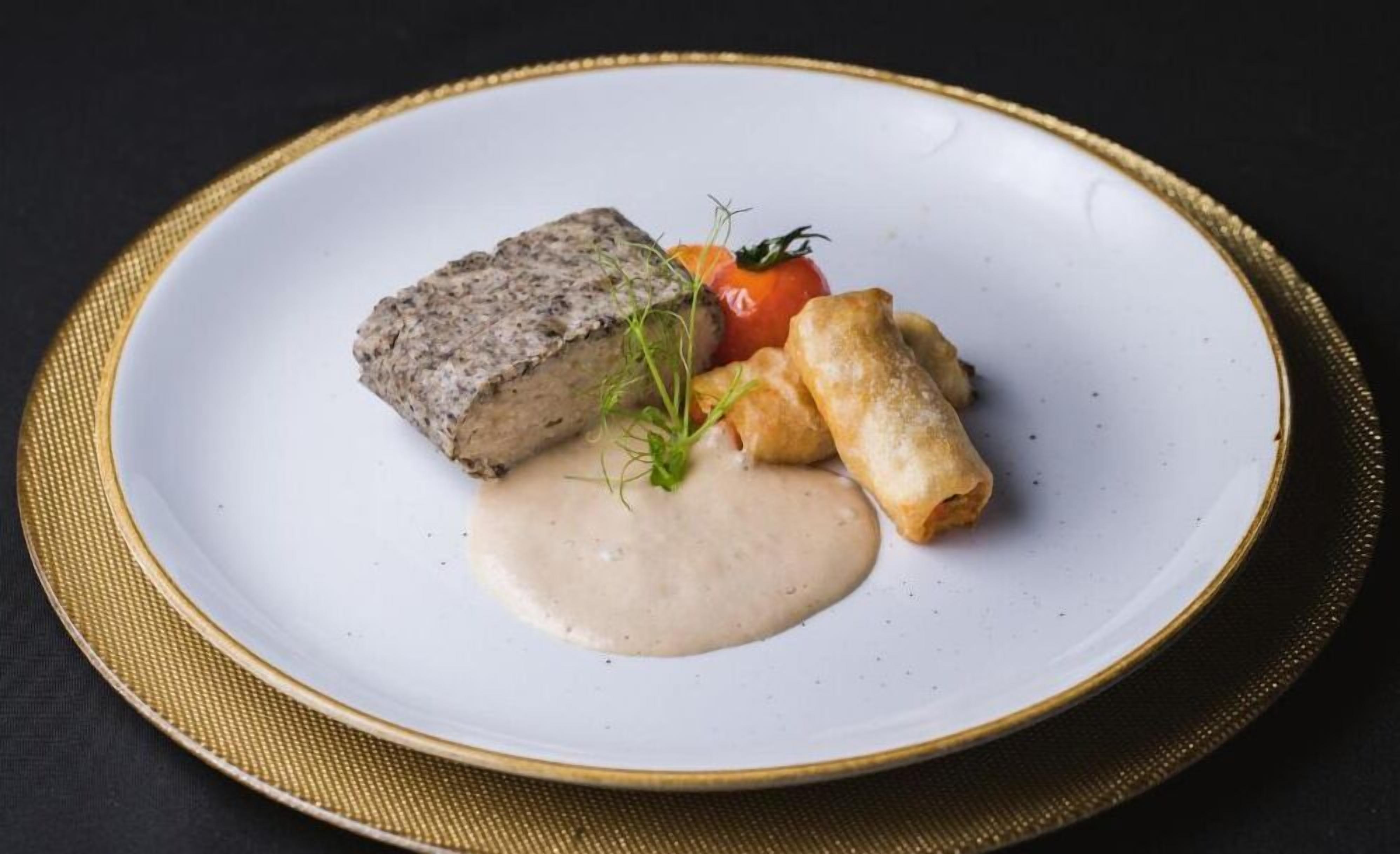 Une assiette blanche ornée d’un bord doré, sur laquelle on voit un morceau de viande, une tomate et deux petits pains.