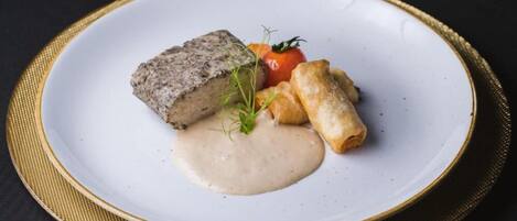 Une assiette blanche ornée d’un bord doré, sur laquelle on voit un morceau de viande, une tomate et deux petits pains.