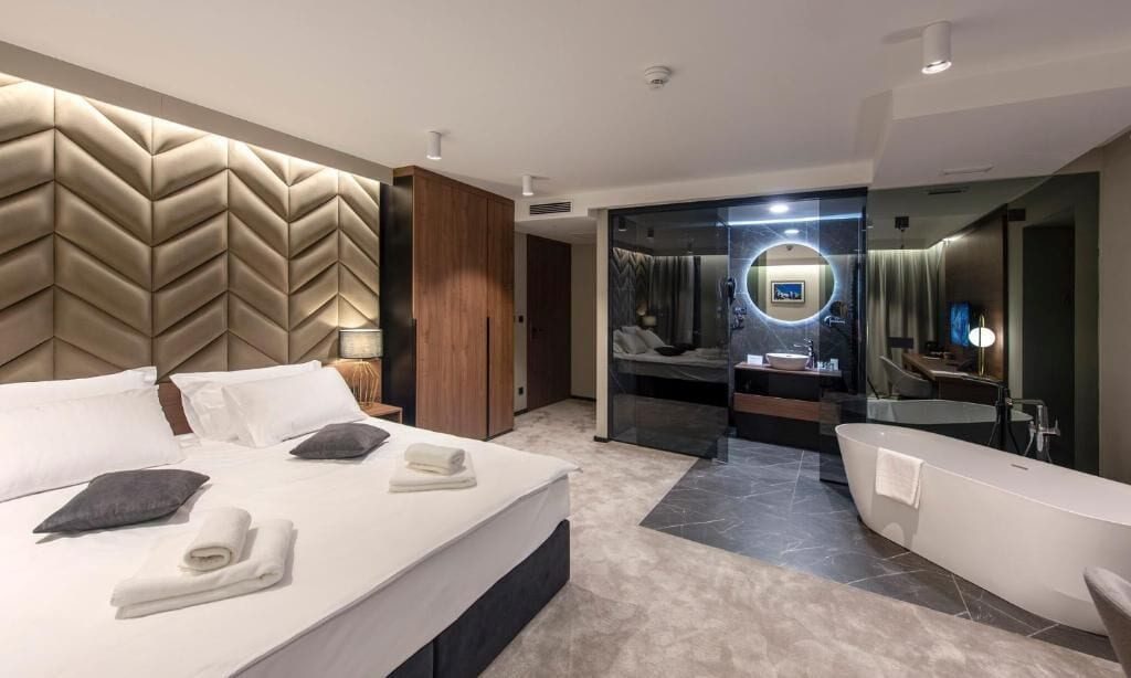 A modern hotel room with a large bed, a separate bathroom with a bathtub, and a mirror with a circular frame.