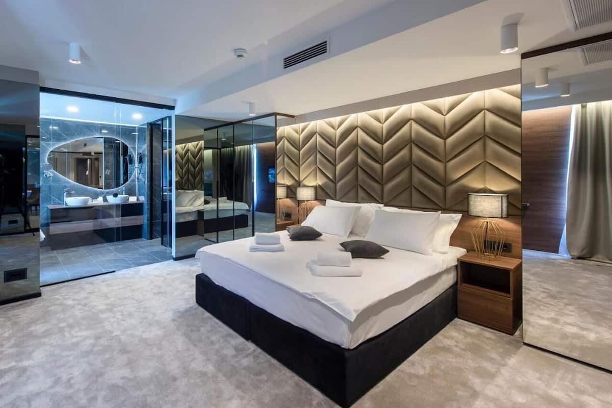 A modern bedroom with a large bed, a glass-enclosed bathroom, and a herringbone-patterned headboard.