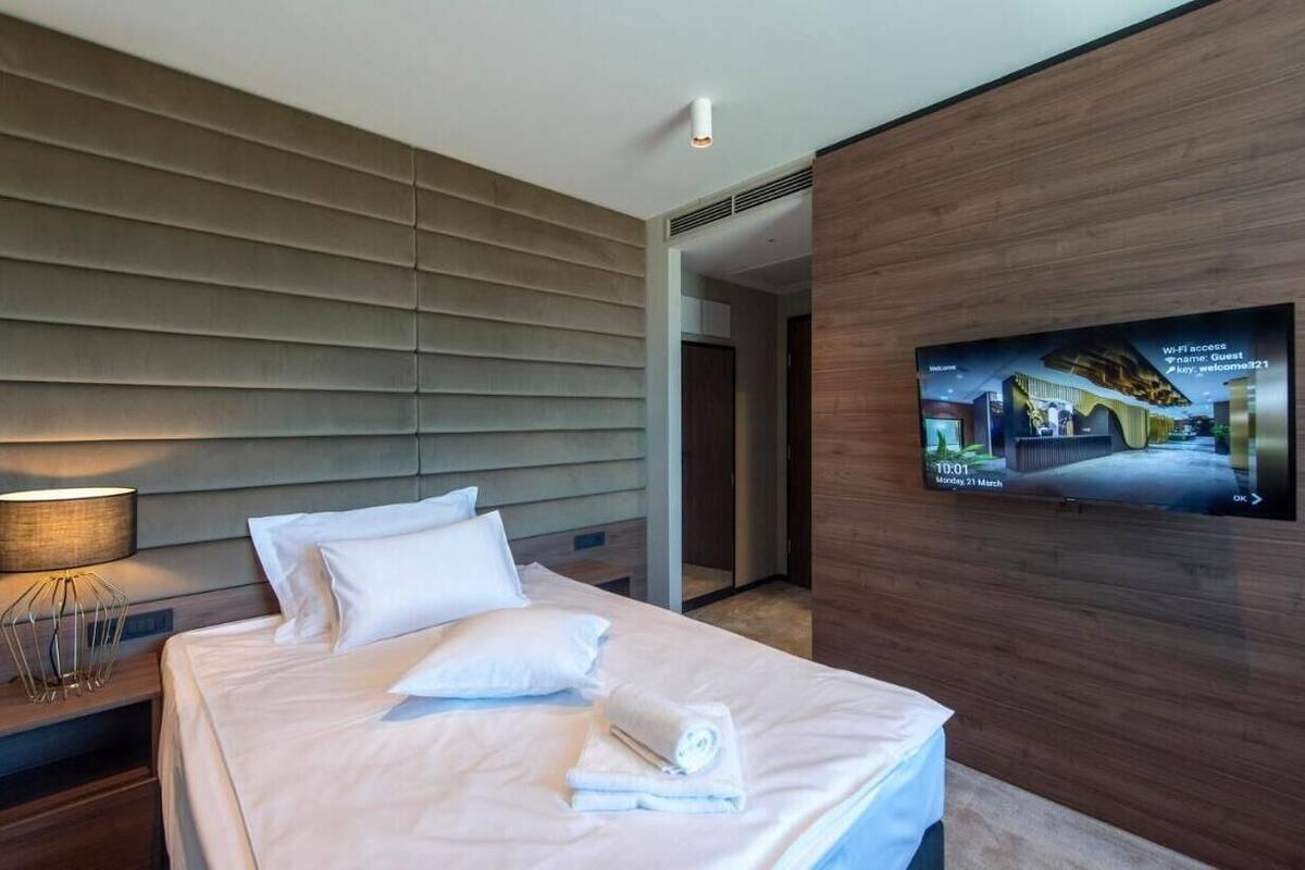 A modern hotel room with a large bed, a flat-screen TV mounted on the wall, and a wooden headboard.