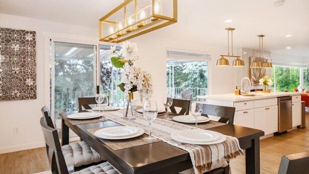 Une cuisine moderne dotée d’un îlot central, de grandes fenêtres et d’un coin repas avec une table en bois foncé, mise pour quatre personnes.