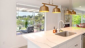 Une cuisine moderne dotée d’un grand îlot central, de luminaires suspendus et offrant une vue sur un balcon aménagé avec du mobilier d’extérieur.