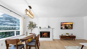 Ein modernes Wohnzimmer mit Kamin, einem Esstisch mit Stühlen, einer Couch und einem großen Fenster mit Blick auf eine verschneite Landschaft.