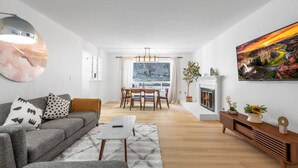 Ein modernes Wohnzimmer mit einem grauen Sofa, einem Couchtisch und einem Essbereich mit Holztisch und Stühlen. Ein Kamin und ein großes Gemälde zieren die Wand.