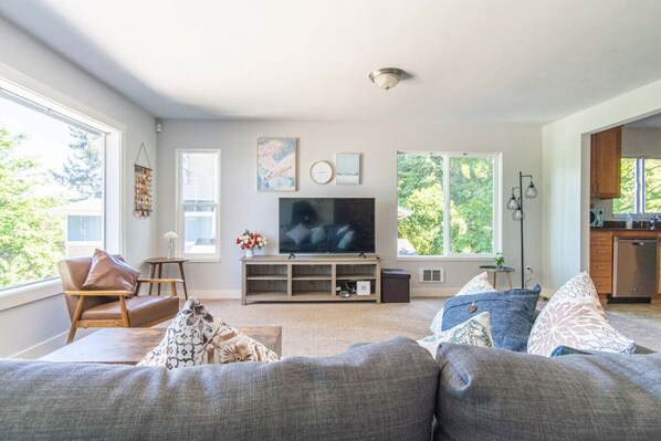 A modern living room with a large flat-screen TV, a sofa, a chair, and a small table with a vase of flowers.