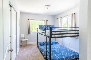 A small, well-lit room with a bunk bed, a small table with a lamp, and a window with curtains.
