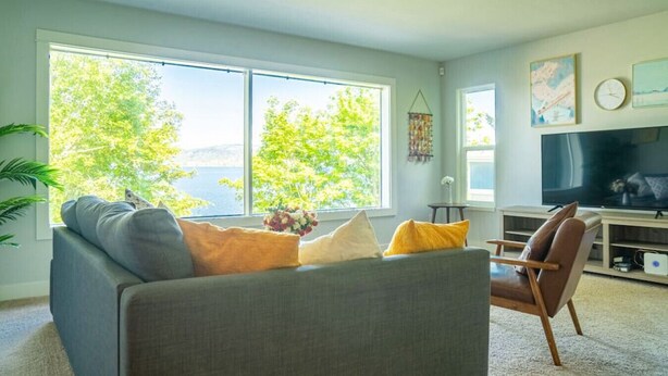 A living room with a sofa, a chair, a TV, and large windows offering a view of trees and a body of water. - 9852 Upper · 4b2b Upper Unit @S Seattle w/ Panoramic Lake View (Seattle)