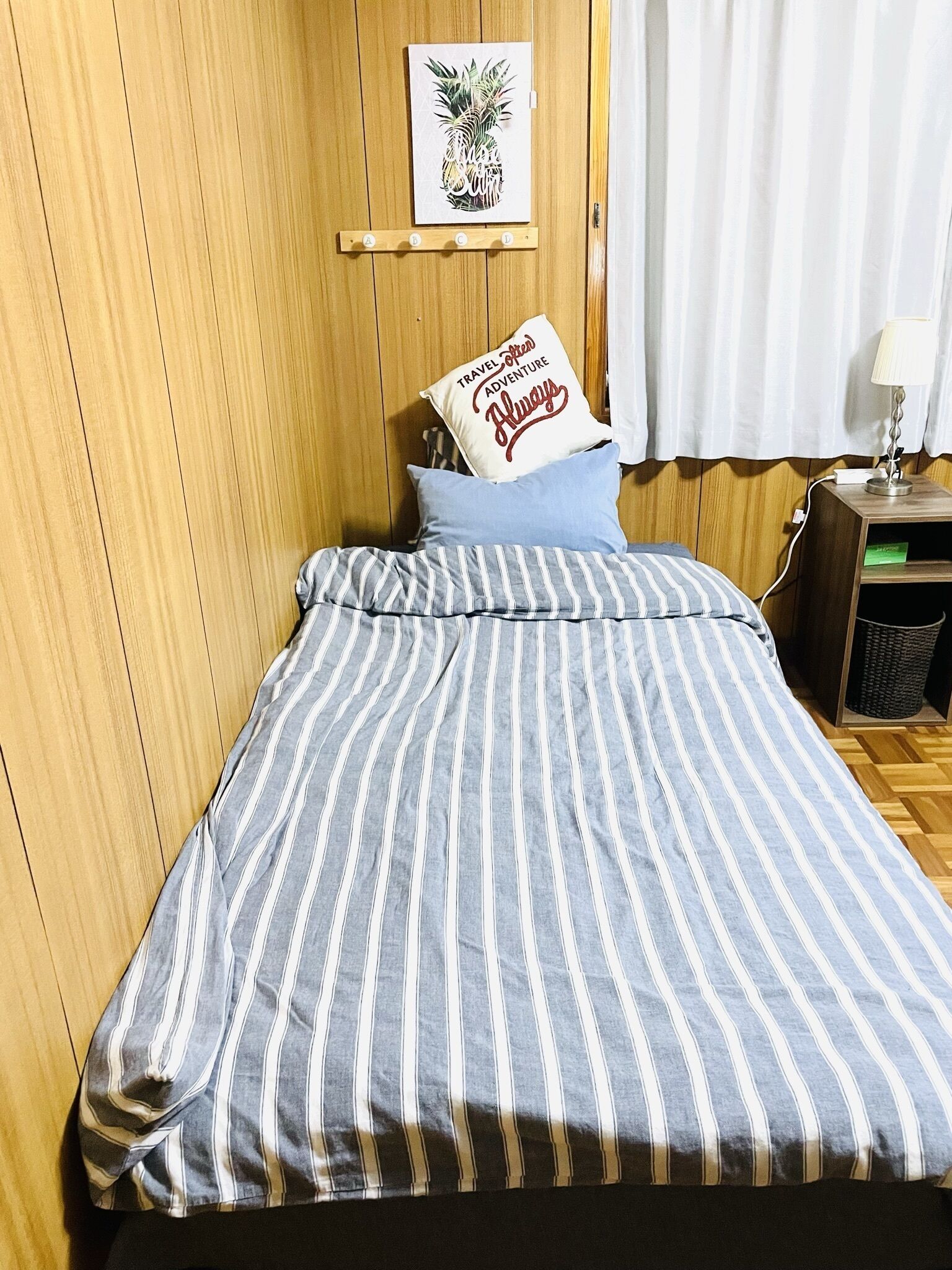 Ein Bett mit gestreifter Bettwäsche, ein Kissen mit der Aufschrift 'Travel Forever Adventure', ein kleines gerahmtes Bild von einer Ananas und ein Nachttisch mit einer Lampe.