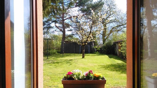 Blick durch ein Fenster auf einen Garten mit einer Holterrasse, einem Rasen und einem Blumenbeet.