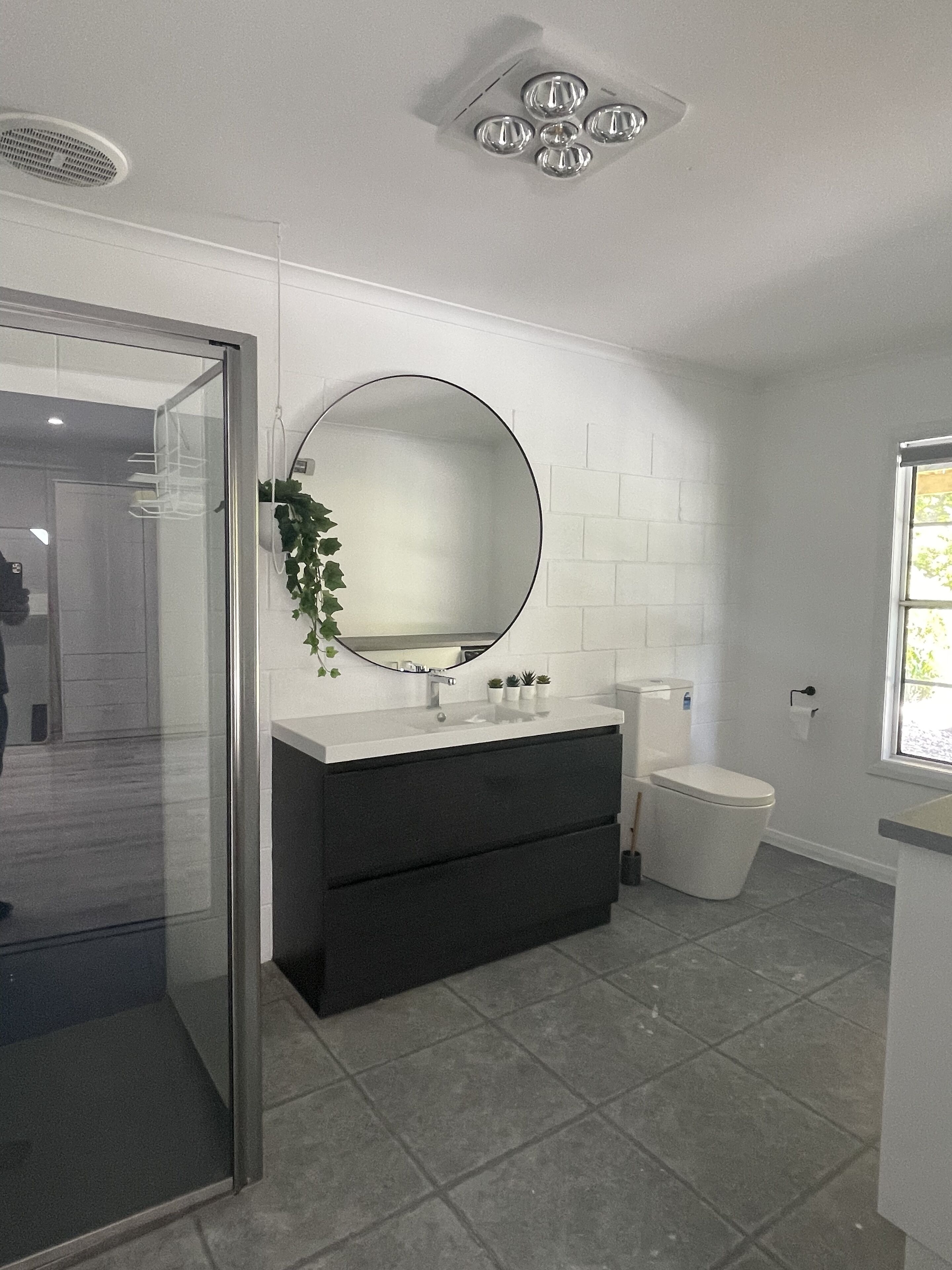 A modern bathroom with a glass shower enclosure, a round mirror, a dark vanity, and a toilet.