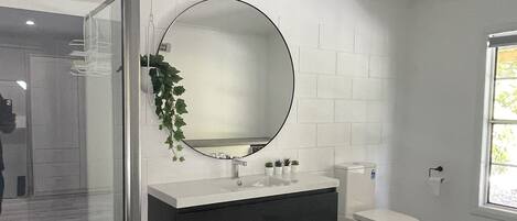 A modern bathroom with a glass shower enclosure, a round mirror, a dark vanity, and a toilet.