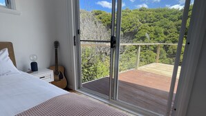 Une chambre avec un grand lit, une guitare et une vue sur un balcon doté d’une terrasse en bois et de verdure.