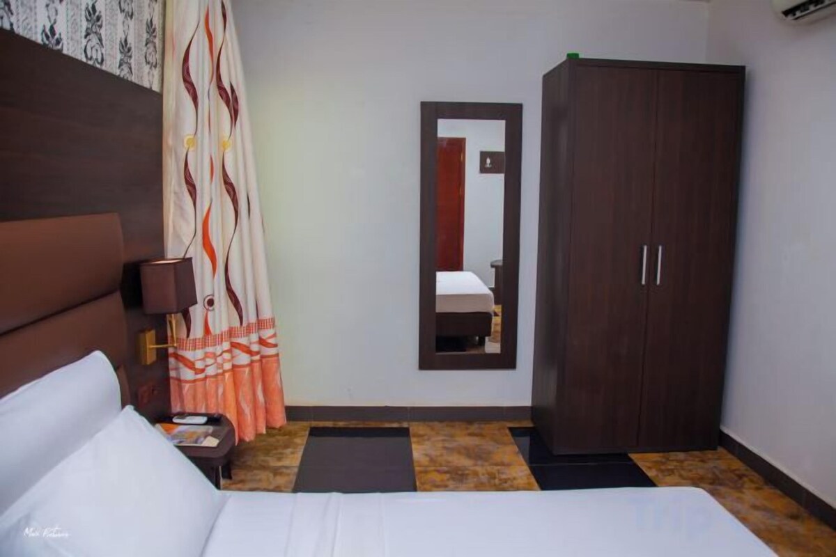 A hotel room with a bed, a wardrobe, a nightstand, a lamp, and a mirror.