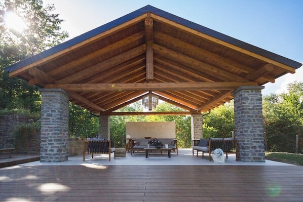 A covered patio with stone pillars, wooden flooring, and outdoor furniture.