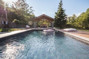 A swimming pool with a wooden gazebo, surrounded by trees and a stone building.