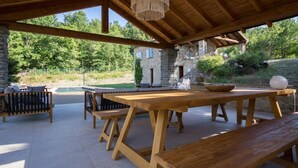 An outdoor patio with wooden tables and benches, a stone building in the background, and a hanging light fixture. - Charming & Authentic Stone House La Brugna (Baselica, frazione di Borgo Val Di Taro)