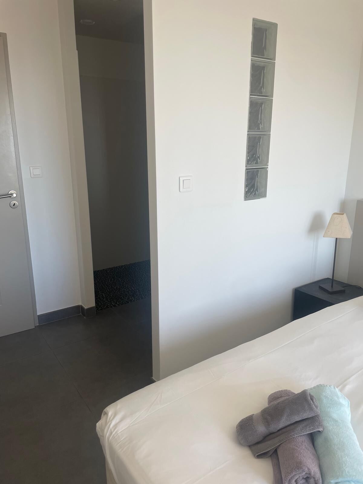 Een hotelkamer met een bed, een nachtkastje met een lamp, een deur en een muur met een lichtschakelaar en een klein raam.