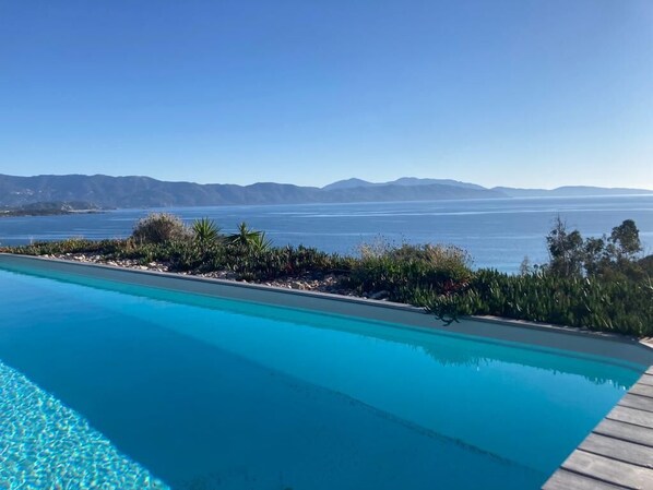 Visão nítida de uma piscina com deck de madeira, rodeada por vegetação ajardinada e com vista para uma vasta extensão de água e montanhas ao longe.