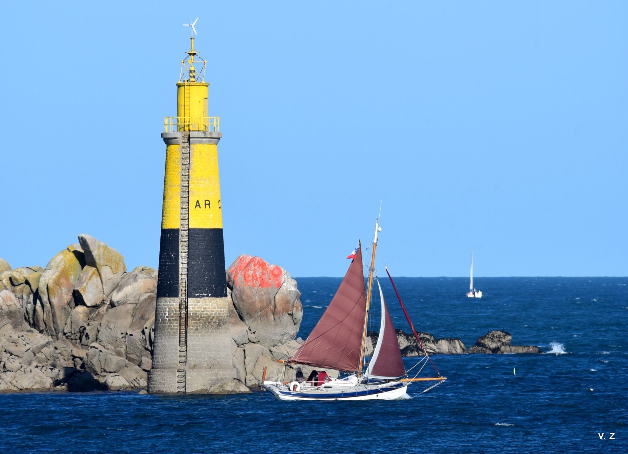 Un phare aux couleurs jaune et noir, un voilier à la voile rouge et un ciel bleu clair.