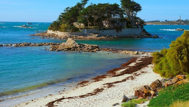 Un paysage côtier avec une île rocheuse couverte d’arbres, des eaux d’un bleu limpide et une plage de sable.
