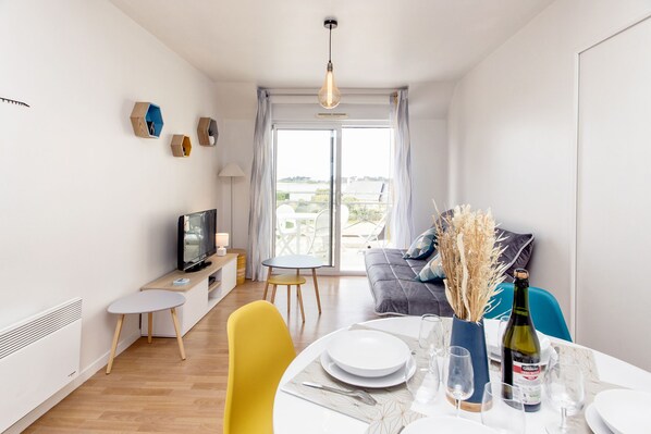 A modern living room with a dining area, a television, and a view through the glass door. - Stunning Apartment with Ocean Views (Roscoff)