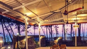 Un restaurante con mesas y sillas de madera, frente a un paisaje de árboles y un atardecer.