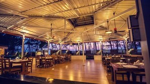 Un restaurante al aire libre con mesas y sillas de madera, techo cubierto y vistas a árboles al anochecer.