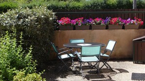 Un espace extérieur aménagé en patio, avec une table ronde et quatre chaises, entouré d’une clôture en bois et agrémenté de diverses plantes.