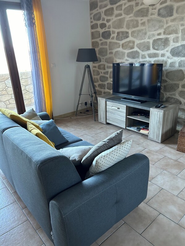 A living room with a sofa, a television on a wooden stand, a lamp, and a stone wall. - High-quality gîte with breathtaking views (LAMA)