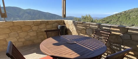 A covered patio with a round table and chairs, stone walls, and a view of distant mountains.
