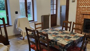 En matplats med ett stort bord täckt av en vacker duk, omgiven av trästolar med röda kuddar. Det finns en tegelkamin på väggen och ett fönster med utsikt över grönskan utanför.