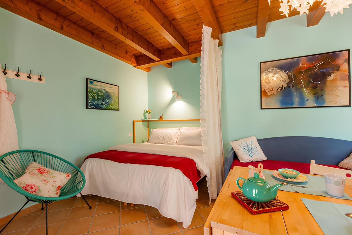 A cozy room with a bed, a chair, a table with a teapot, and a painting on the wall.