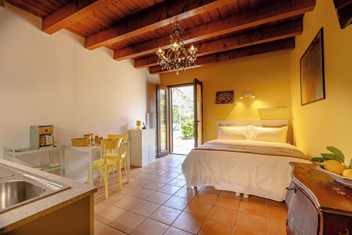 A bedroom with a bed, a chandelier, a dining area with chairs, and a kitchen with a sink.