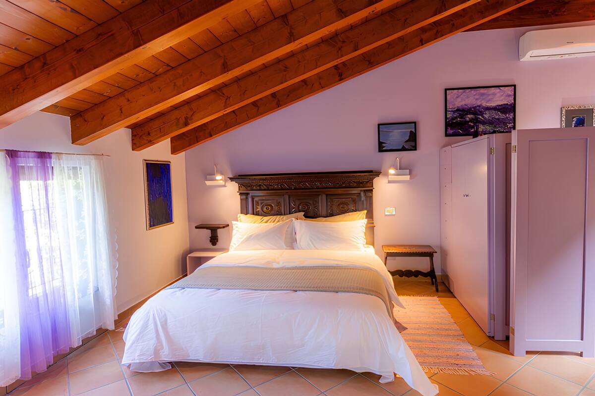 A bedroom with a wooden ceiling, a bed, a nightstand, a wall-mounted TV, and a refrigerator.