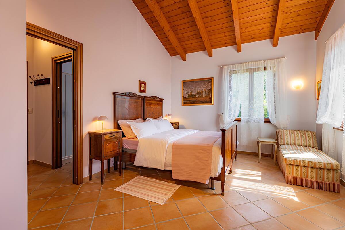 A bedroom with a wooden ceiling, a bed with white bedding, a nightstand, a chair, and a window with curtains.