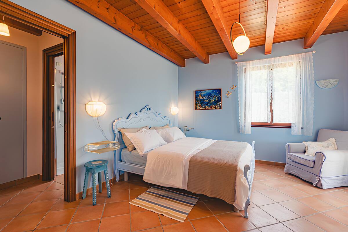 A bedroom with a wooden ceiling, a bed with white bedding, a nightstand, a chair, and a window with curtains.