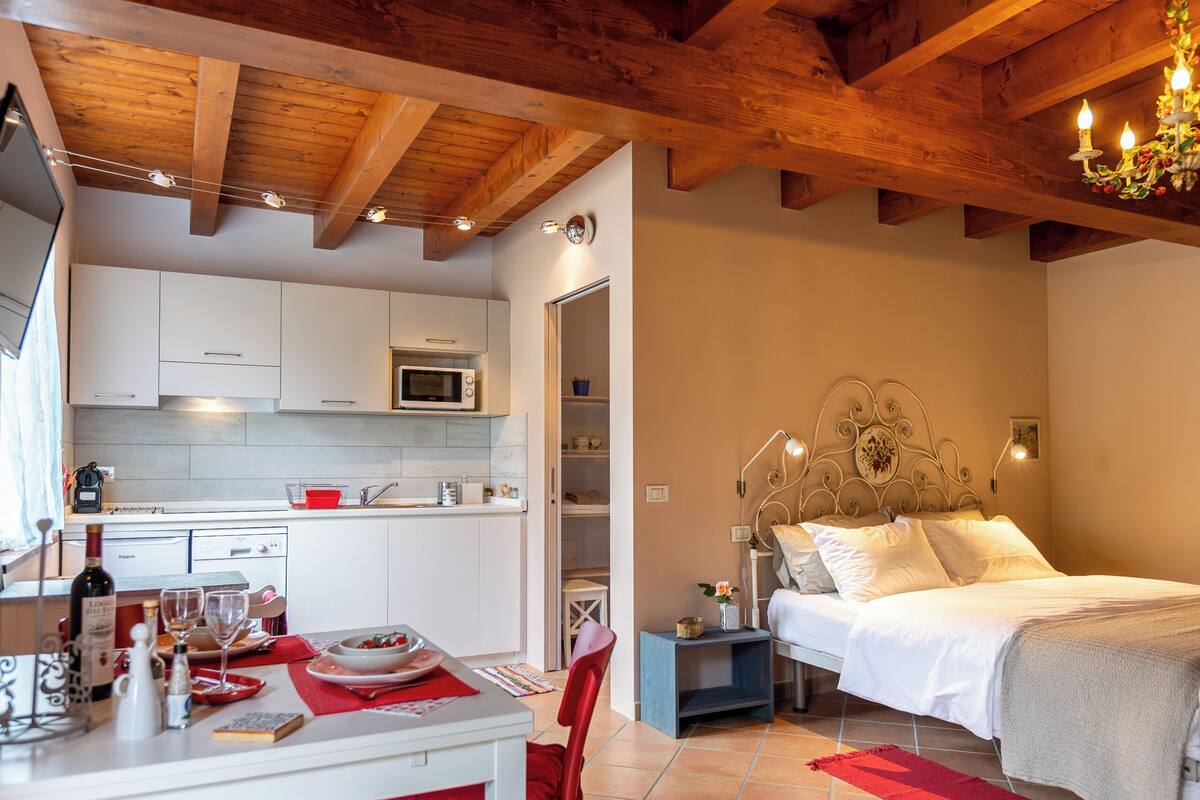 A modern bedroom with a bed, bedside table, and a small bedside lamp. Adjacent is a kitchen with white cabinets, a microwave, and a red countertop.