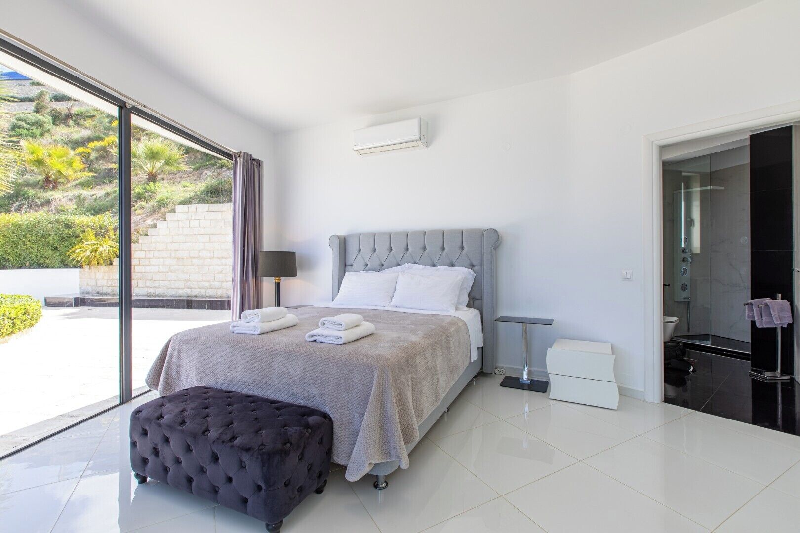 Une chambre moderne avec un grand lit, une table de chevet, un petit tabouret et une salle de bain équipée d’une douche et de toilettes.