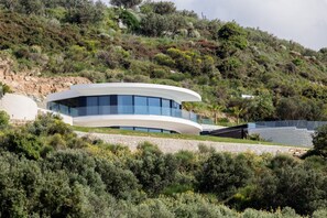 Un bâtiment moderne aux lignes courbes, doté de grandes fenêtres, entouré d’une végétation luxuriante et adossé à une colline rocheuse.
