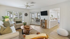 Un salon spacieux comprenant un canapé, un fauteuil et une table ronde, un téléviseur fixé au mur, et un balcon visible à travers les portes vitrées.
