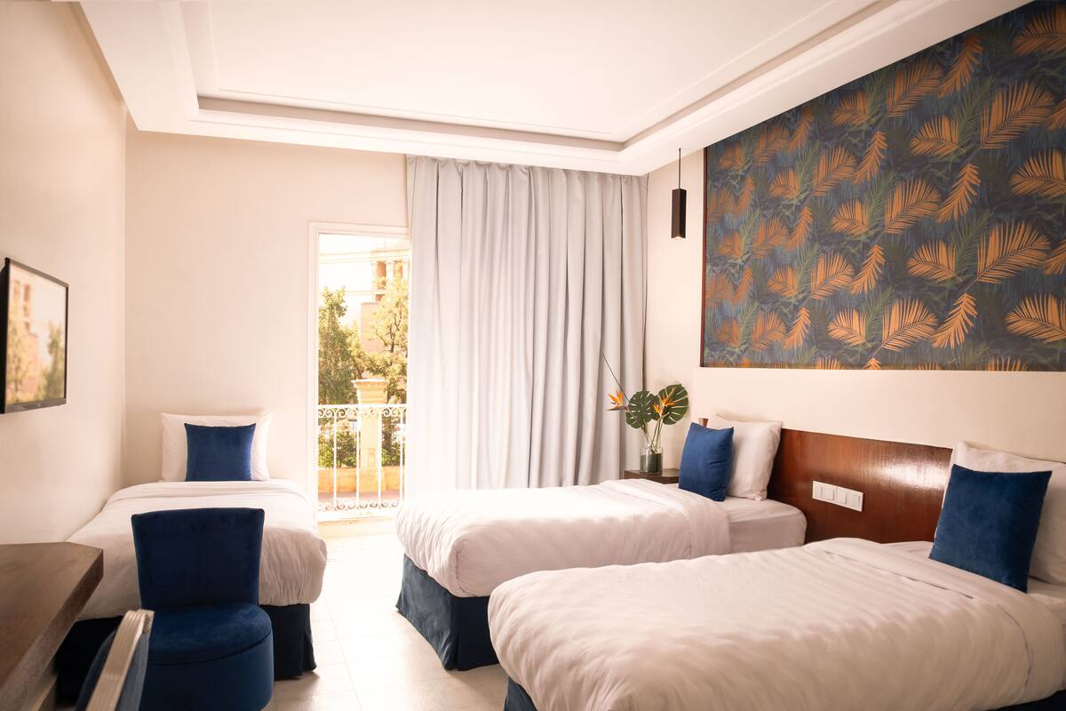 A hotel room with two beds, a large window with curtains, a painting on the wall, and a balcony with a view.